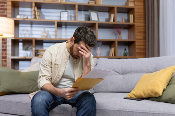 istock Upset man at home sitting on sofa reading letter with bad news notification in envelope opening, unhappy in living room and sad 1473957796