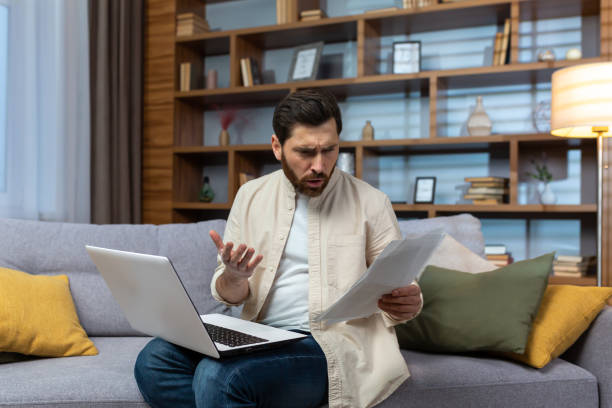 istock A worried young man is sitting on the sofa at home, holding documents and a laptop. Problems with accounts, rent, business, credit, loan 1473958603