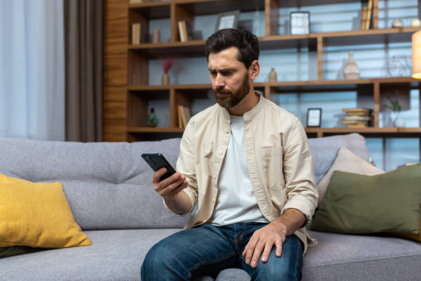 istock Communication problems. A serious young man in a beige shirt sits on the sofa at home, holds the phone, looks worriedly at the screen, types a message 1473960697