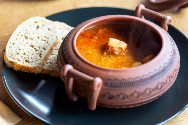Pot of hot bograch soup and two slices of bread on black plate. Restaurant serving. Selective focus, defocus