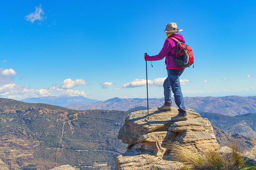 A woman on top of a rock, with backpack and hiking poles, looking towards the horizon and enjoying the impressive landscape of mountains and valleys at her feet, surrounded by nature and fresh air