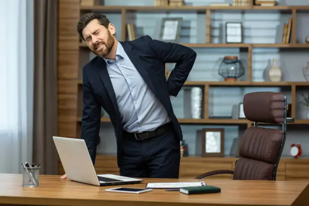 Photo of The problem of sedentary work. A young businessman man in a suit stands in the office at the desk and holds his back. Feels severe pain and spasms, massages with his hand, grimaced from pain