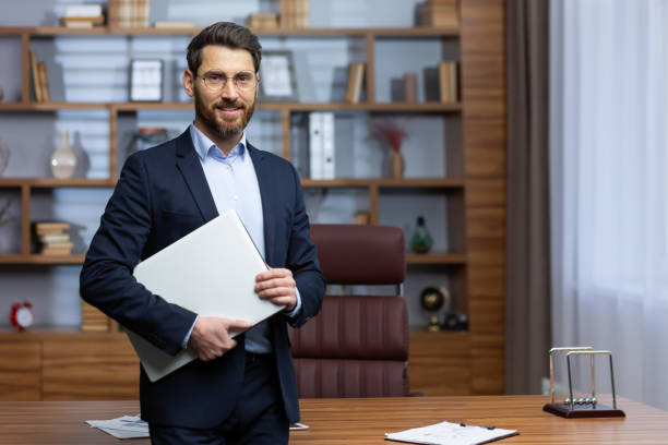 portrait of mature businessman inside office, senior man in business suit and beard standing and holding laptop, boss at work smiling and looking at camera - lawyer young adult suit expressing positivity imagens e fotografias de stock