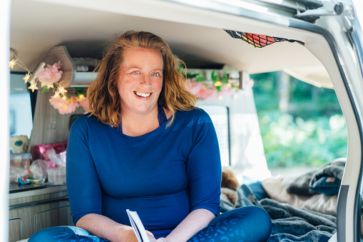 Portrait of smiling middle-aged woman relaxing, reading book while sitting in camper car vehicle during road trip. Enjoying free lifestyle, travel, vacation, freedom. Selective focus.