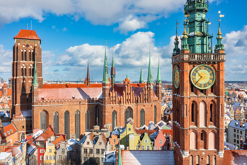 large panorama showcases the vibrant cityscape of Gdansk, Poland, on a sunny day with a clear blue sky. The view, captured from the viewpoint of the Forum shopping mall, encompasses the old city with notable landmarks such as Wyżynna Gate (Brama Wyżynna) and Długouliczna Gate (Brama Długouliczna) in the frame. The image depicts a bustling street filled with cars and various forms of public transport. Pedestrian areas are teeming with people, adding a sense of movement and liveliness to the scene.