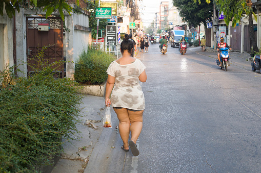 Rear view of walking young thai woman in dress  with mobile phone. Woman is walking  in residential district of Bangkok Chatuchak. In her hand she has a bag with food, In street some people are walking or driving motorcycle.