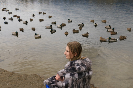 A Ukrainian/Russian model next to a pond with lots of ducks. She is wearing long blond straight hair, and a black and white pattern coat.