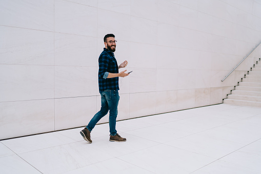 Full length portrait of cheerful hipster guy in spectacles for vision correction and earphones listening positive music songs during leisure daytime, happy Caucasian man with mobile technology