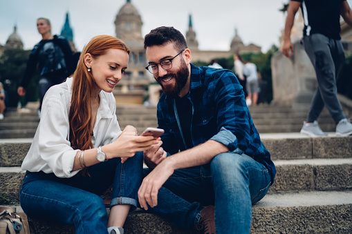 Cheerful boyfriend and girlfriend watching positive travel vlog during cellular networking in city, funny Caucasian hipster guys enjoying together journey and smartphone messaging with best friends