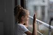 Little sad girl pensive looking through the window glass with a lot of raindrops. Sadness and loneliness childhood concept image.