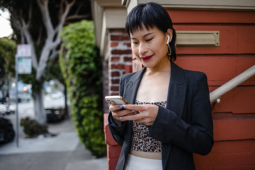 A modern woman in stylish clothes smiling and looking at her smart phone. A woman with in-ear headphones is texting online.