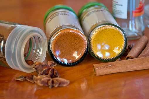 A close-up of a red chilli spice bottle and yellow turmeric spice bottle surrounded by star anise and cinnamon sticks on a wooden kitchen top background.