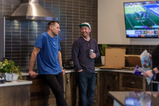 Two mid adult men wearing casual clothing enjoy talking and watching a football game on TV while standing in the kitchen during a casual social gathering with family and friends.