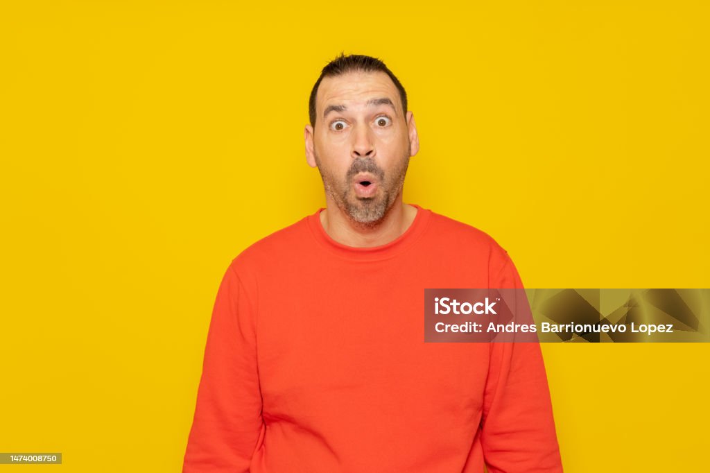 Hispanic man with a beard in a red sweatshirt with his eyes wide with emotion, his mouth is open in surprise generated by something unheard of. Isolated on yellow background. Hispanic man with a beard in a red sweatshirt with his eyes wide with emotion, his mouth is open in surprise generated by something unheard of. Isolated on yellow background Cheering Stock Photo