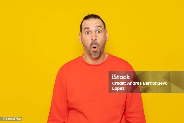 Hispanic Man With A Beard In A Red Sweatshirt With His Eyes Wide With Emotion His Mouth Is Open In Surprise Generated By Something Unheard Of Isolated On Yellow Background Stock Photo - Download Image Now