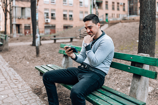 A young Caucasian man is sitting on a bench and looking at his phone.