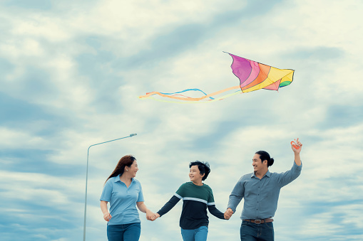 Progressive happy family vacation and carefree day concept. Young parents mother father and son run along and flying kite together road with enjoy natural scenic on scenery and clear sky background.