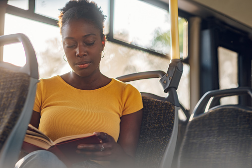 Young adorable african american joyful woman sitting in a bus and reading a book. Public transportation and people concept. Copy space. Diverse portrait.