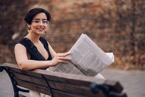 Portrait of a beautiful smiling female in stylish office clothes sitting on a bench and reading newspaper with a brick wall in the background. Business woman style. Fashion photo.