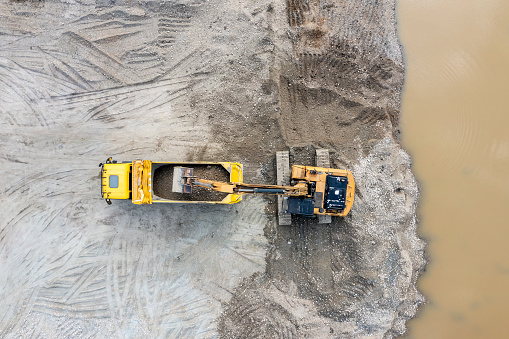 Sand extraction for the construction industry. The excavator extracts sand from the lake and loads it onto the dump truck, aerial view.