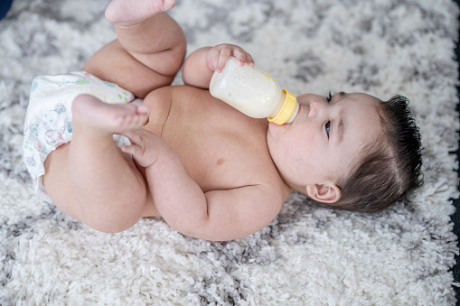 A sweet little baby boy of Hispanic decent, lays on a rug as he feeds himself a bottle.  He is wearing only a diaper and appears very content.