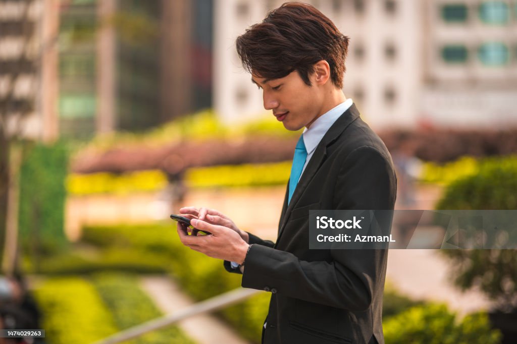 Young Asian Male Adult Using his Cellphone Waist up shot of young asian male adult wearing a dark suit and a sky blue tie. He is using his cellphone in the middle of a public park. 20-24 Years Stock Photo