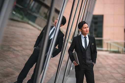 Outdoor business portrait of confident man wearing a black suit and holding a notebook. Attractive asian man standing next to modern building and looking away.