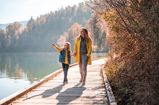 Autumn walk of a beautiful Caucasian mother and her daughter, holding hands and discussing something the girl is pointing at. Full length image, wide shot, reflection on the lake next to women.