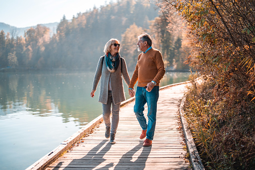 A couple of Caucasian seniors enjoying a walk on a lakefront in a forest, full of autumn colors. Full length image, looking at each other. Partial shadow of them on the path in front.