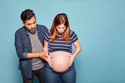 Adult latin couple surprised feeling prenatal contractions, isolated on blue background. Woman about to give birth. Concept of due date. Studio photo. Copy space.