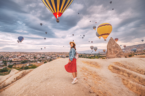 Sunlight aglow on her dress, happy young woman stands in the countryside of Cappadocia, the silhouettes of hot air balloons dancing in the sky above.