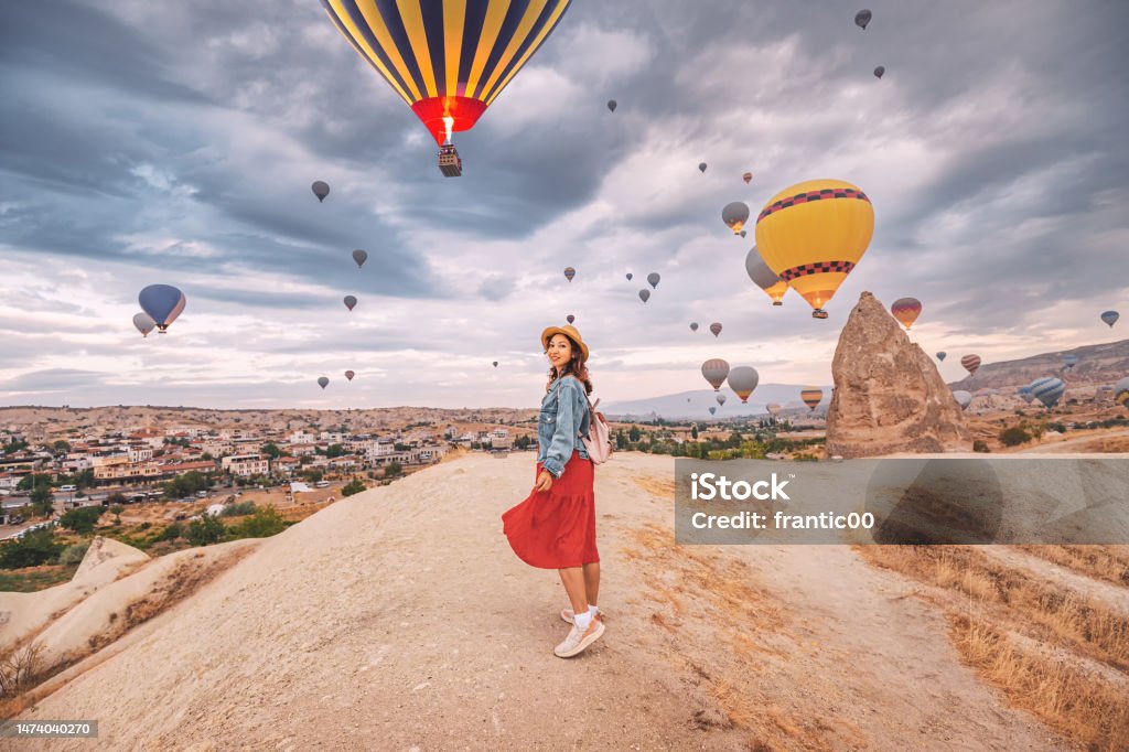 Sunlight aglow on her dress, happy young woman stands in the countryside of Cappadocia, the silhouettes of hot air balloons dancing in the sky above. Cappadocia Stock Photo