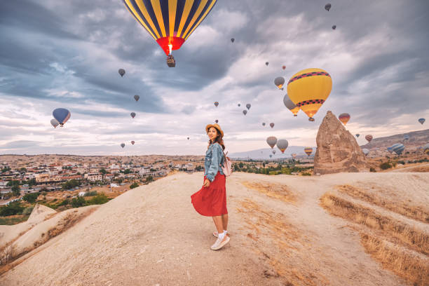 Sunlight aglow on her dress, happy young woman stands in the countryside of Cappadocia, the silhouettes of hot air balloons dancing in the sky above. stock photo