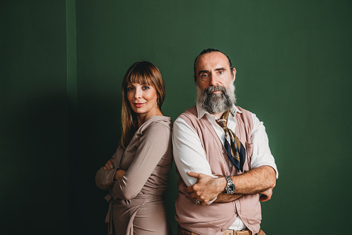 Portrait of a mature adult couple against a green wall. They are wearing elegant clothes.