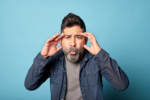 Mid latin adult man surprised in denim t-shirt, hands on face isolated on blue background. Confused male stunned, shocked by unexpected news or information astonished. Shock content. Copy space.