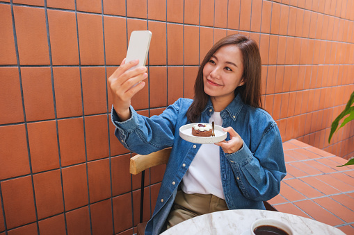 Portrait image of a young woman using mobile phone to take a selfie with a cake in cafe