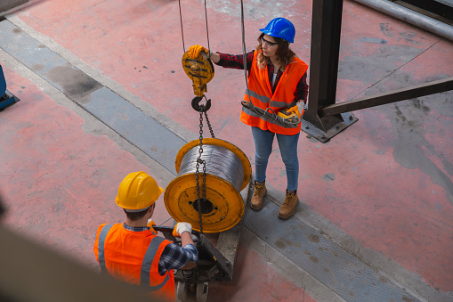 Young heavy industry worker man and engineer woman holding crane hook button working with safety workwear and moving a massive metal construction object in warehouse of factory.