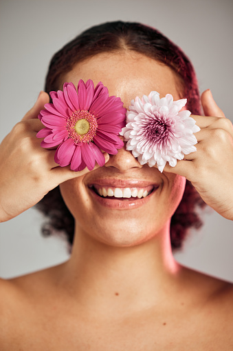 Woman, face and flower eyes with smile for natural skincare, beauty or cosmetics isolated on a grey background. Happy female, person or lady smiling in happiness holding colorful plant petals for art