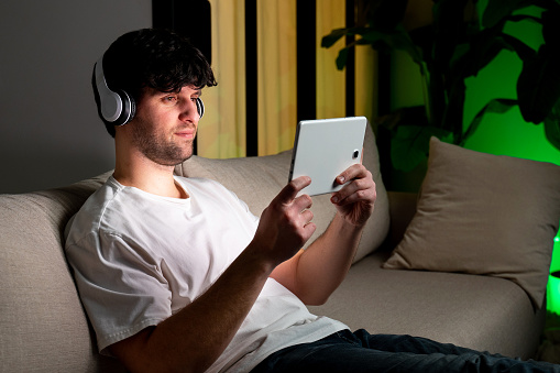 Young man relaxing on sofa in his living room. he is listening to a music on headphones and using digital tablet.