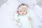 portrait of a happy and smiling little baby girl in a crib on a white cotton bed with a bow on her head with blue eyes and a green bodysuit
