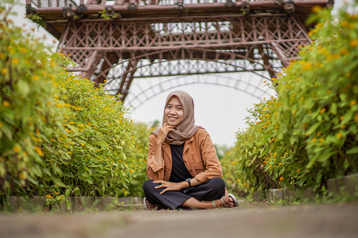 beautiful asian muslim woman in modern hijab clothes sitting in the middle of a flower garden road. flower garden with replica eiffel tower