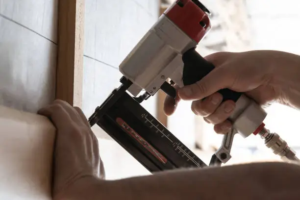 Close up unrecognizable male hands using pneumatic nailer, stapler gun for wooden plank outdoors. Clipping with metal nails and staples, making layout on wooden timber. Build house, carpenter job