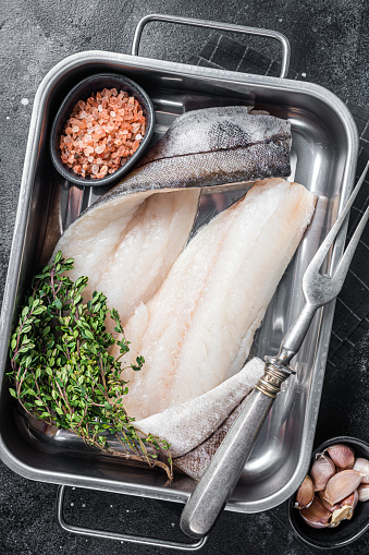 Raw haddock fish fillets, whitefish meat in kitchen tray with thyme. Black background. Top view.