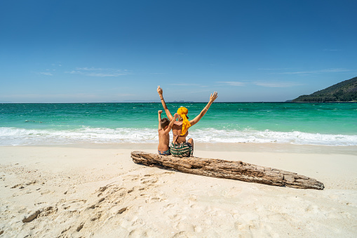 Happy woman having fun with son on a tropical beach, enjoying summer holidays, raising hands up. Tourism, family travel concept. Real people lifestyle.