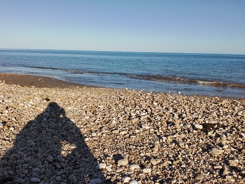The main object of this photo is a beach with lots of pebbles on the beach and there is a shadow of someone sitting on the beach
