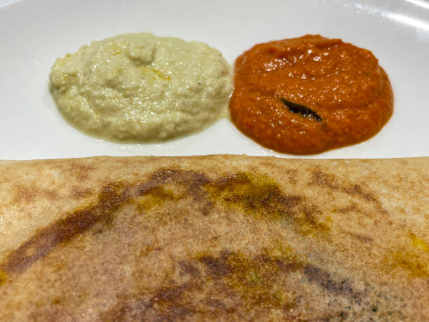 istock Close up image of Indian breakfast masala dosa cut open, thin folded potato pancake served on white plate with dips / condiments / chutneys, including sambar, elevated overhead view 1474115056