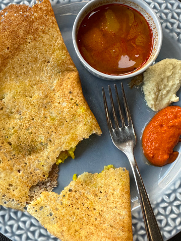 Stock photo of a breakfast meal with an Indian masala dosa, a thin potato pancake that is folded and filled, it is being served on a blue plate with a selection of dips / condiments / chutneys, including sambar
