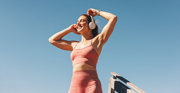 Caucasian sports woman listening to music on headphones outdoors. Woman in sportswear standing against the sky. Happy female athlete taking a break from her workout.