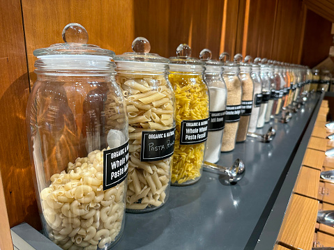Stock photo showing pantry with an assortment of glass jars full of various dried ingredients, including pasta, beans, seeds, lentils, pulses and spices. These jars are lined up and organised neatly for ease of access, with labels on them.