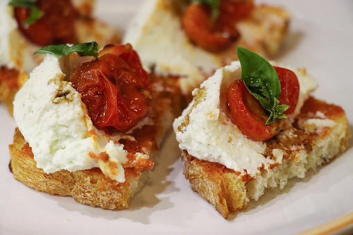 Stock photo showing close-up view of bruschetta recipe open sandwich with baguette bread slices, garlic and basic pesto sauce, chopped cherry tomatoes on mozzarella, drizzled with balsamic vinegar and garnished with fresh basil leaves on white plate.
